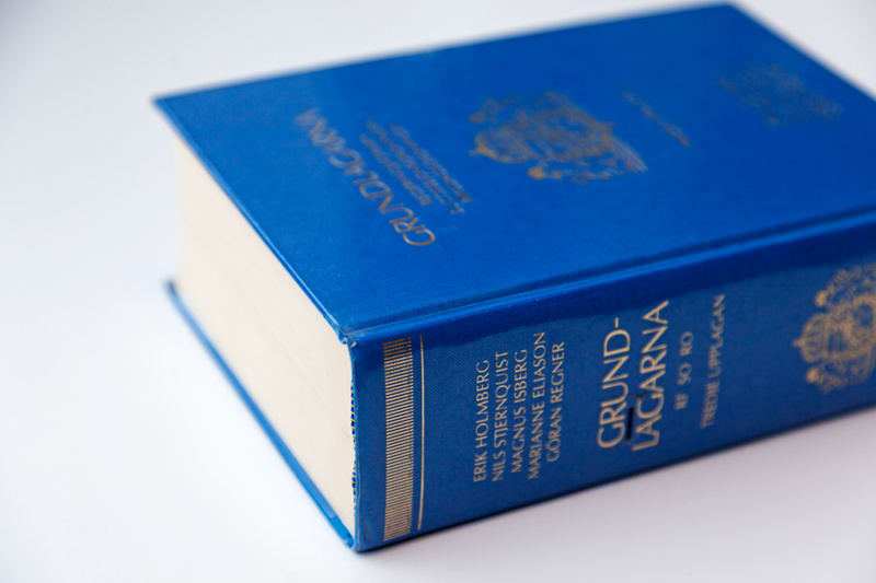 En närbild av en tjock, blå inbunden bok som ligger på en vit yta. Titel och författare är tryckta i guldfärg på bokens framsida och rygg. På framsidan syns texten "GRUNDLAGARNA" samt en symbol, troligtvis ett vapen, också i guldfärg. På bokryggen står "GRUNDLAGARNA", ”RF SO RO” och ”TREDJE UPPLAGAN”, följt av namnen ERIK HOLMBERG, NILS STJERNQUIST, MAGNUS ISBERG, MARIANNE ELIASON och GÖRAN REGNER. Boken verkar vara en lagbok eller en referensbok. Bilden är tagen med ett grunt skärpedjup där boken är i fokus medan bakgrunden är oskarp. Belysningen är jämn och vit.