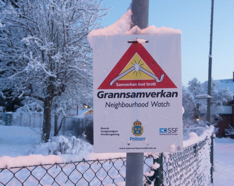 En vertikal bild visar en skylt för 'Grannsamverkan' (Neighbourhood Watch) monterad på en stolpe, sett mot en snötäckt bakgrund. Skylten är vit med en stor röd triangel överst som innehåller ett symboliskt motiv av en ljusstråle som träffar en bruten