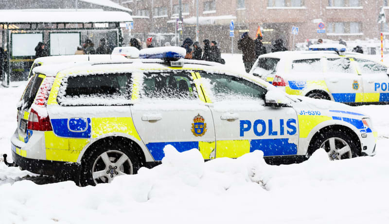 Bilden visar två svenska polisbilar, främst kombimodeller, parkerade utomhus under kraftigt snöfall. Den främre bilen är en vit polisbil med blå och gula reflexdekaler, med texten "POLIS" tydligt synlig på sidan samt Polisens emblem. Taket och delar