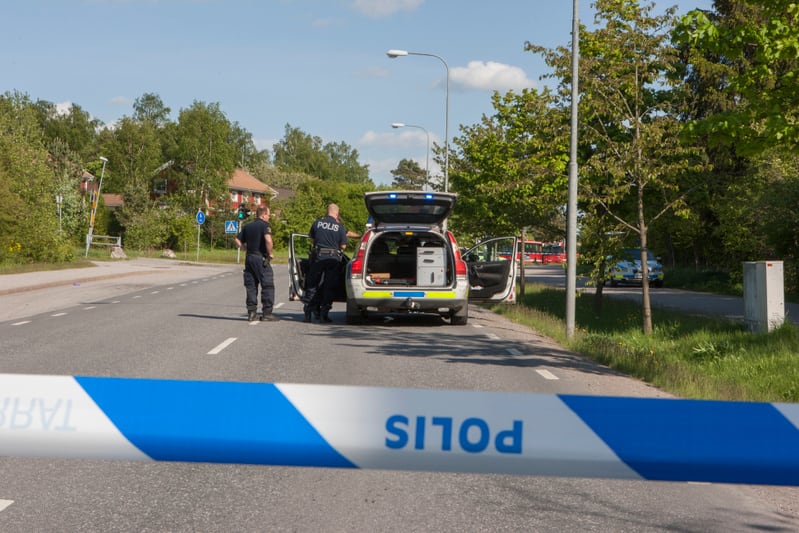 Bilden visar två polismän vid en svensk polisbil som står parkerad på sidan av en väg. Polisbilens baklucka och en sidodörr är öppna, och den har tänd blå belysning. En blåvit avspärrningstejp med texten "POLIS" löper i förgrunden. I bakgrunden syns