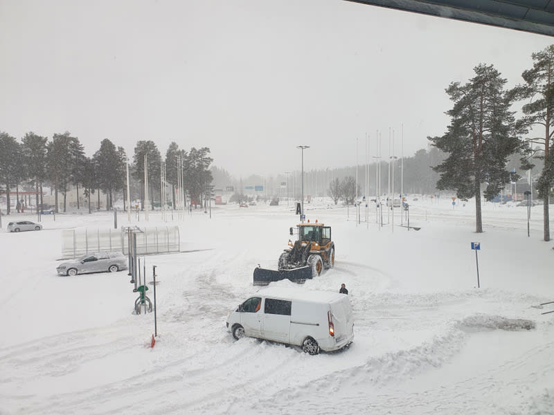 En bild visar en vinterdag med snöfall över ett utomhusområde. I förgrunden syns en vit skåpbil som delvis är täckt av snö och en gul hjullastare som plogar snö. En person står nära hjullastaren. Parkerade bilar, även de delvis snötäckta, syns i bakg