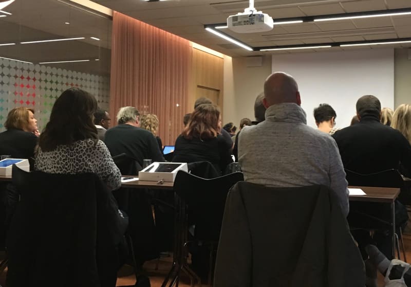 Bilden visar en grupp människor som sitter på stolar vid bord, vända mot en projektionsyta i ett rum. Ett flertal personer ses bakifrån i förgrunden, några med kort hår och andra med längre hår. På borden framför dem syns vita fyrkantiga etikettmaski