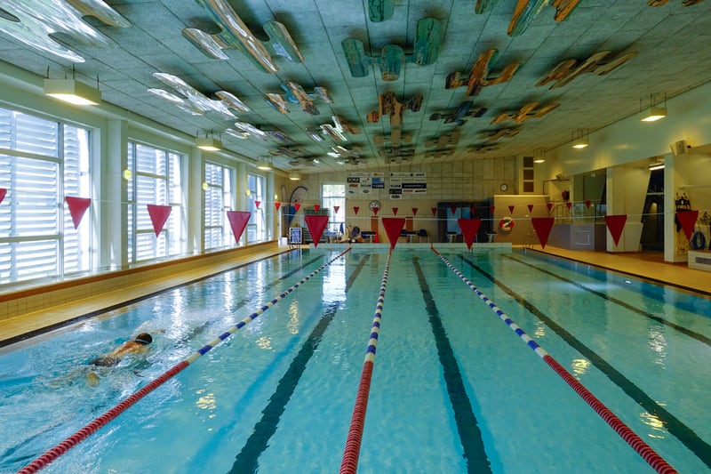 Bilden visar en inomhuspool med en person som simmar i förgrunden. Poolen har tydliga banmarkeringar med linor i rött, vitt och blått. Taket ovanför poolen är prytt med flera träliknande figurer som liknar flygplan. Längs ena långsidan av poolen finn
