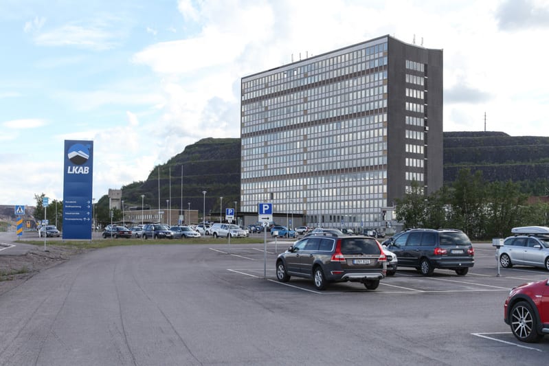Bilden visar ett stort parkeringsområde med flera bilar parkerar, en stor hög byggnad med många fönster, samt en stor, blå skylt med texten LKAB och pilar som pekar mot olika avdelningar som "Huvudentré", "Besöksparkering", "Industriområde" och "Pers