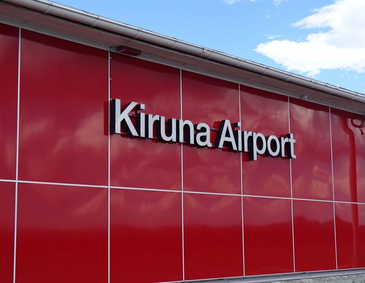 Bilden visar fasaden på en byggnad med en skylt där det står "Kiruna Airport". Fasaden är täckt av röda paneler med vita eller ljusgrå linjer som bildar ett rutmönster. Bokstäverna på skylten är vita med ett mörkt djup som skapar en 3D-effekt. Ovanfö