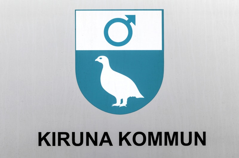 Bilden visar Kiruna kommuns vapen och namnet "KIRUNA KOMMUN" skrivet med svarta versaler undertill. Vapnet är en sköld uppdelad horisontellt i två fält. Det övre fältet är vitt med en manssymbol (Mars-symbol) i blått. Det nedre fältet är blått och vi