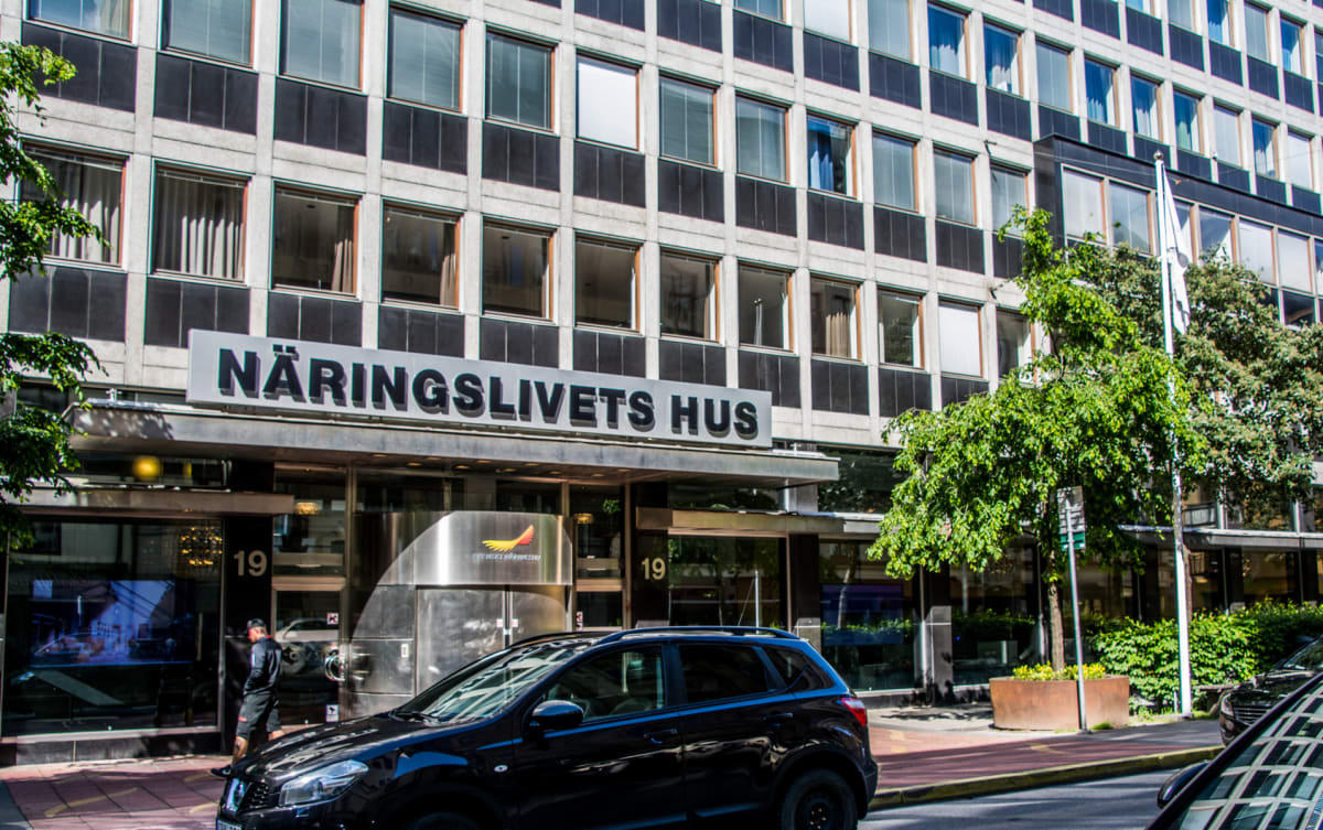 Bilden visar fasaden på en större byggnad med texten "NÄRINGSLIVETS HUS" ovanför ingången. Byggnaden har många fönster med persienner och en modern, rutig design med grå och svarta paneler. Framför byggnaden syns ett grönt träd och en svart bil parke