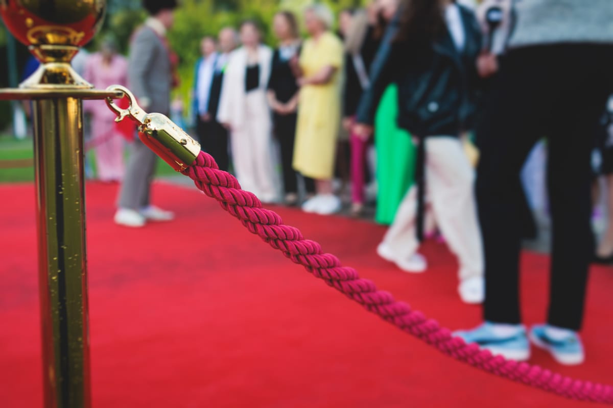 Bilden visar en guldglänsande avspärrningsstolpe med ett rött rep som sträcker sig horisontellt över bildytan. Repet är tvinnat och fäst vid stolpen med en guldpläterad krok. Under repet och stolpen ligger en röd matta. I bakgrunden syns suddiga männ