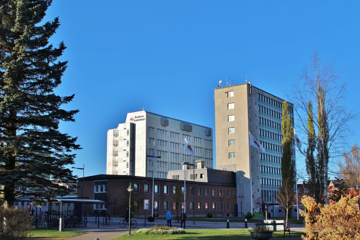 Bilden visar flera byggnader i vad som framstår som en stadsmiljö under dagtid. Till vänster syns delar av ett stort barrträd. I mitten och till höger domineras bilden av höga byggnader, varav en har texten "Bodens Kommun" och en logotyp på takkanten