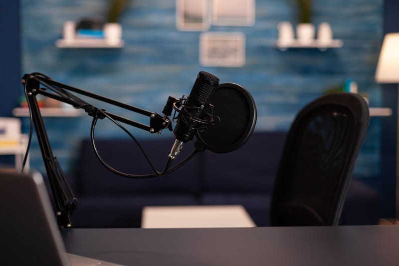 Bilden visar ett mörkt rum, troligtvis en studio eller ett hemmakontor. I förgrunden till vänster syns delar av en silverfärgad laptop på en mörk bordsskiva. Mot mitten och höger förgrund står en studiemikrofon monterad på en mikrofonarm, utrustad med ett platt popfilter. Till höger om mikrofonen syns baksidan av en svart kontorsstol med ryggstöd i nät. I bakgrunden syns en vägg med en blå texturerad tapet eller målad yta. Två vita hyllplan med olika föremål är monterade på väggen. En mörk soffa skymtar också i bakgrunden. En golvlampa med vit skärm syns i bildens högra kant.