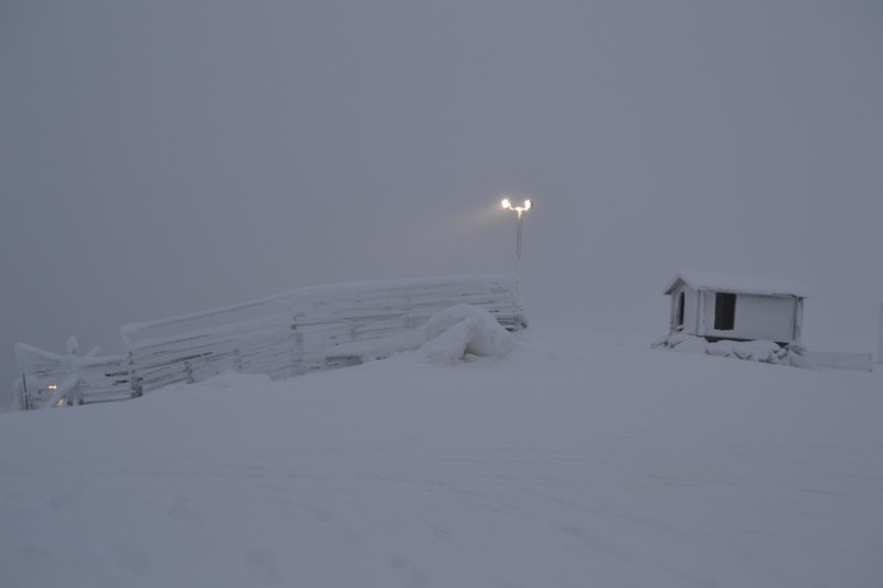 Bilden visar ett snötäckt landskap under en vinterdag eller natt. I förgrunden syns ett stort område täckt av snö. På höger sida av bilden finns en liten byggnad helt täckt av snö. Till vänster om byggnaden, i mitten av bilden, står en gatlykta med t