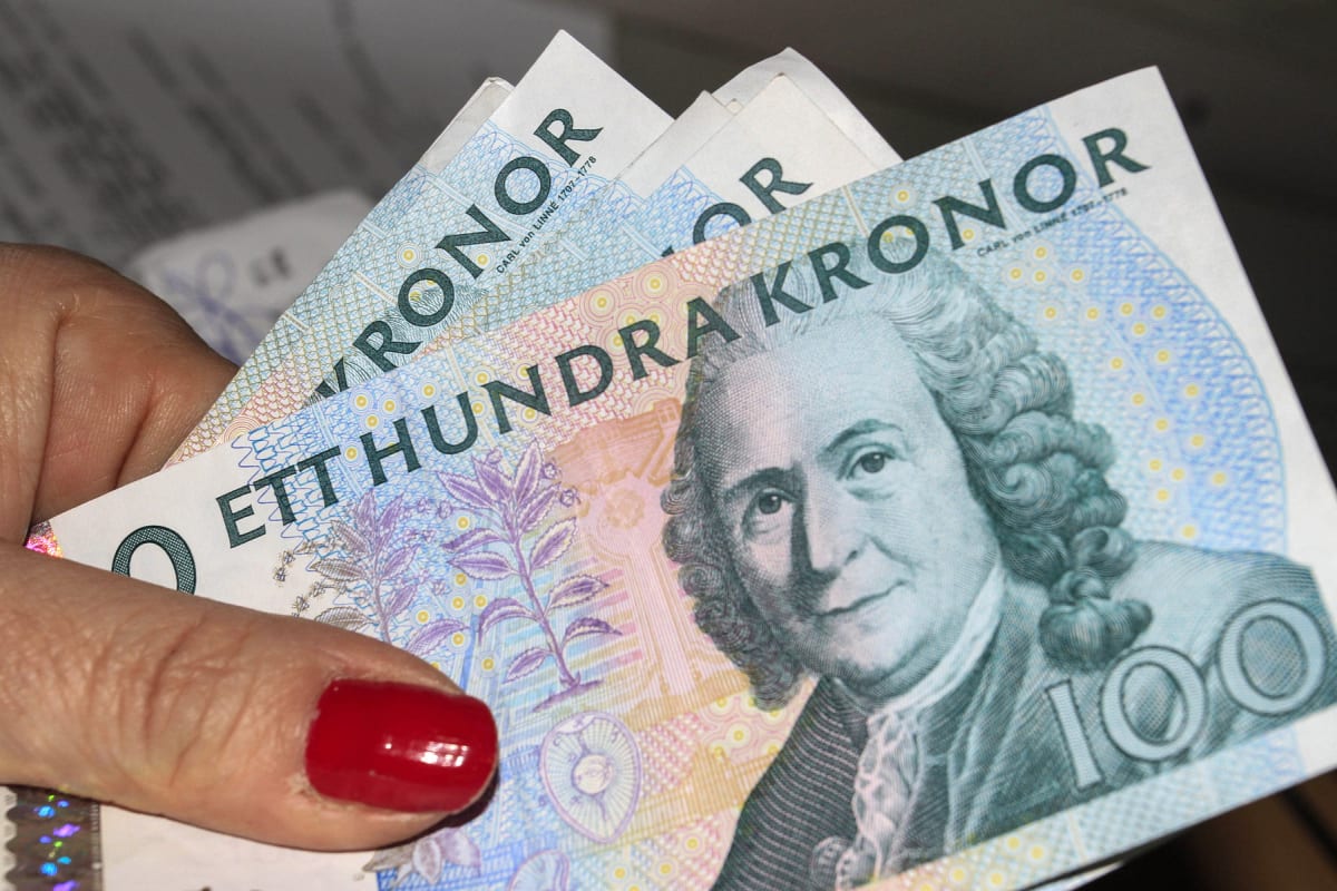 En hand med rött nagellack håller upp flera svenska hundrakronorssedlar. Sedlarna är vita med färgade detaljer i blått, lila och rosa. Framträdande på sedlarna syns texten "ETTHUNDRA KRONOR" och ett porträtt av en man med peruk, som är Carl von Linné