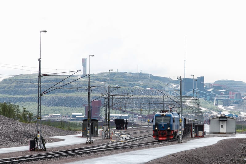 En vidvinkelbild tagen utomhus vid en gruvanläggning eller industri. I förgrunden syns ett järnvägsspår med en blåfärgad lokomotiv med röda detaljer och ett antal tågvagnar. Längs spåret finns flera elledningsmaster med överliggande ledningar samt be