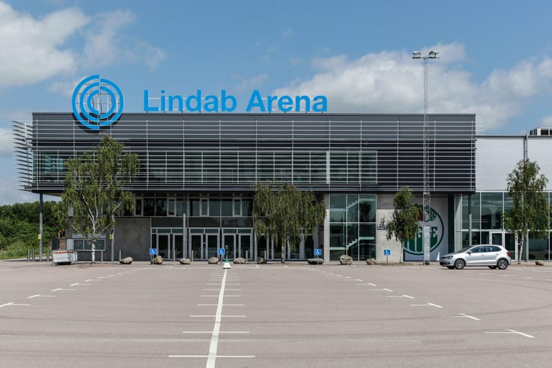 Bilden visar fasaden på en modern byggnad med ett stort parkeringsområde framför. Byggnaden har horisontella lameller på ovandelen och stora glaspartier med dörrar på undervåningen. Ovanför taklinjen syns en blå logotyp och texten "Lindab Arena". På