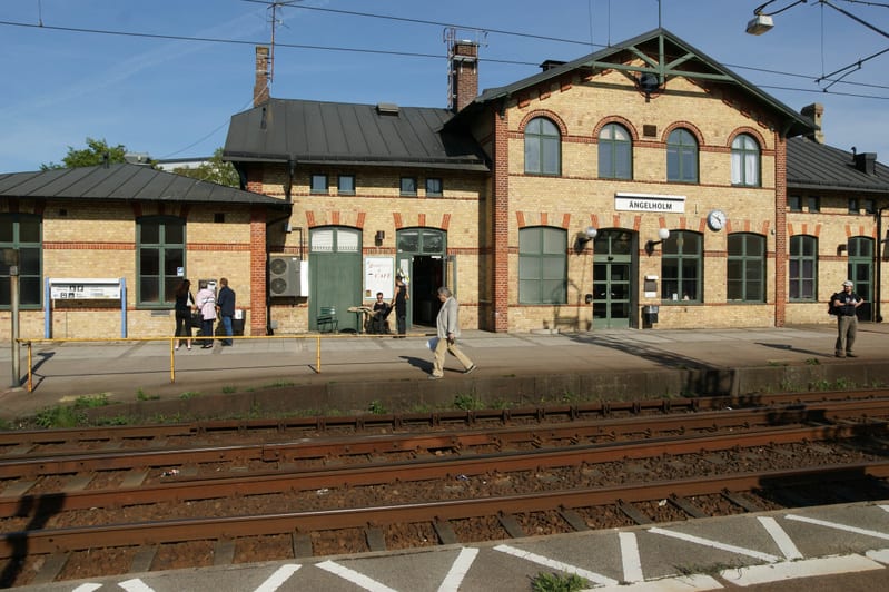 En bild tagen i dagsljus som visar en tågstation med ett gult tegelhus som huvudfokus. På husets fasad syns namnet "ÄNGELHOLM" ovanför en av ingångarna. Byggnaden har flera fönster med bågformade överdelar och gröna fönsterkarmar. Utanför byggnaden f