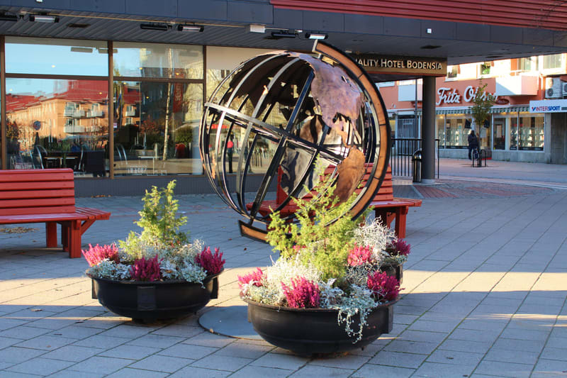 Bilden visar ett torg eller en öppen plats framför en byggnad med stora fönsterpartier. I förgrunden finns två stora svarta blomlådor med växter som inkluderar gröna buskar och lila blommor. Bakom blomlådorna står två röda bänkar. Mellan bänkarna och