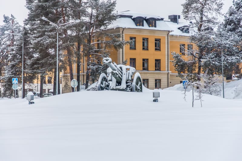 Bilden visar ett snöigt vinterlandskap med en stor, äldre kanon täckt av snö i förgrunden. Bakom kanonen står en stor gul byggnad med många fönster och ett tak täckt av snö. Runt byggnaden och kanonen finns flera snötäckta barrträd. Det ligger tjocka