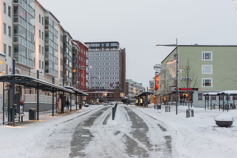 En gata täckt av snö och slask med byggnader på båda sidor under ett grått, molnigt vinterlandskap. På vänster sida syns vita och röda byggnader, varav några har balkonger med glasräcken. Längs gatan finns flera busshållplatser med tak och bänkar. Mi