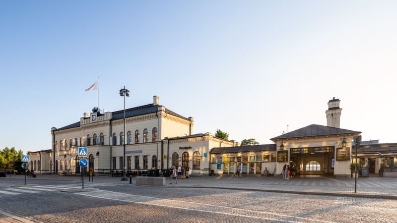 En vidvinkelbild tagen utomhus under dagtid, som visar en stor, ljusgul byggnad med många fönster och mörka tak, som förmodligen är en tågstation. Byggnaden har detaljer som en flaggstång med en vit flagga och en klocka högst upp. Framför byggnaden f