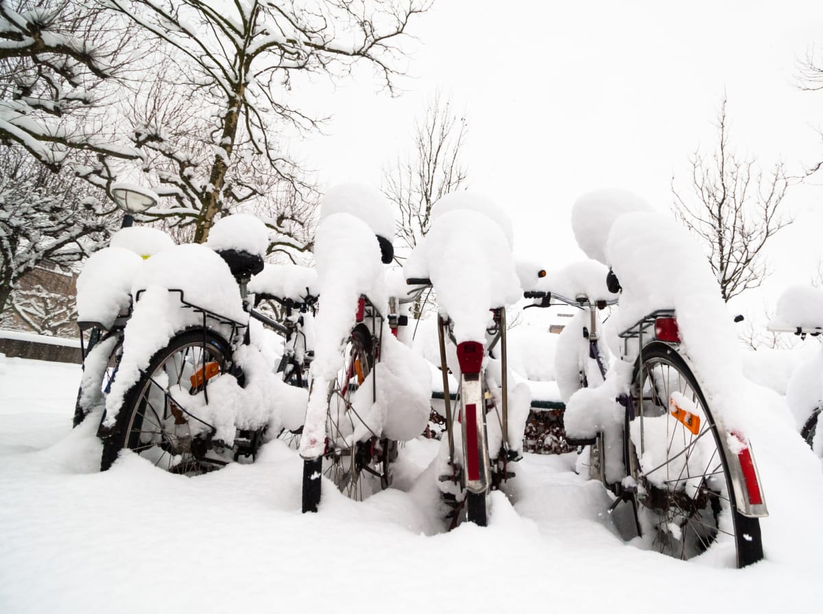 Bilden visar ett snötäckt utomhusområde där flera cyklar står parkerade. Markytan är helt täckt av ett tjockt lager snö. Cyklarna är också rikligt täckta av snö, särskilt på sadlar, pakethållare och styren, vilket gör att endast delar av deras ramar,