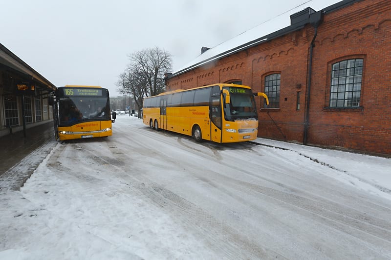 En bild som visar en snöig utomhusmiljö med två gula bussar. Till vänster syns delvis en buss med destinationen "Trelleborg via Svedala" och linjenummer "165" på displayen. Bussens registreringsskylt är av svensk modell. Vägen och gångbanan är täckt