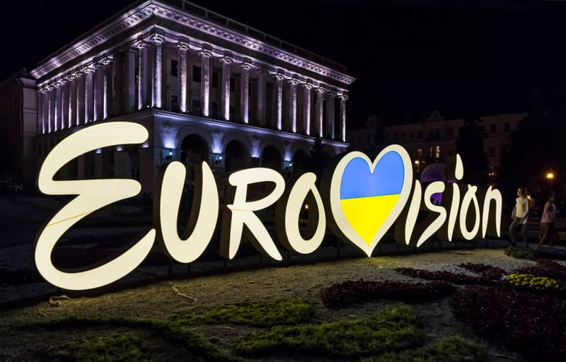 Bilden visar en nattlig scen med det stora, belysta ordet "Eurovision" i förgrunden, där bokstaven 'V' är ersatt med ett hjärta som innehåller den ukrainska flaggans färger, blått och gult. Bokstäverna är ljust färgade och lyser upp den mörka omgivningen. Mellan bokstäverna och under dem syns vegetation, inklusive gräs och rödaktiga blomsterarrangemang. I bakgrunden reser sig en stor byggnad med pelare längs fasaden, upplyst av lila och vita strålkastare som framhäver dess arkitektur. Ett par otydliga personer ses ståendes bredvid "Eurovision"-skylten på höger sida. Hela scenen är badad i ett mörkt nattljus, med de upplysta elementen som fokuspunkter.