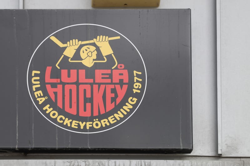 Bilden visar en närbild på en svart rektangulär skylt, troligen av metall, monterad på en ljusgrå vägg. I mitten av skylten finns en cirkulär logotyp med en vit ram. Inuti logotypen, på en svart bakgrund, syns ordet "LULEÅ" i rött, stort typsnitt, med "HOCKEY" i ännu större rött typsnitt direkt under. Runt den nedre halvan av cirkeln står texten "LULEÅ HOCKEYFÖRENING 1977" i guldfärgat typsnitt. Ovanför "LULEÅ HOCKEY" finns en grafisk bild i guldfärg föreställande en person i hjälm som håller en ishockeyklubba. Skylten har synliga spår av smuts eller vattenfläckar. Den övre delen av bilden visar en del av en gulaktig kvadratisk struktur som är delvis synlig.
