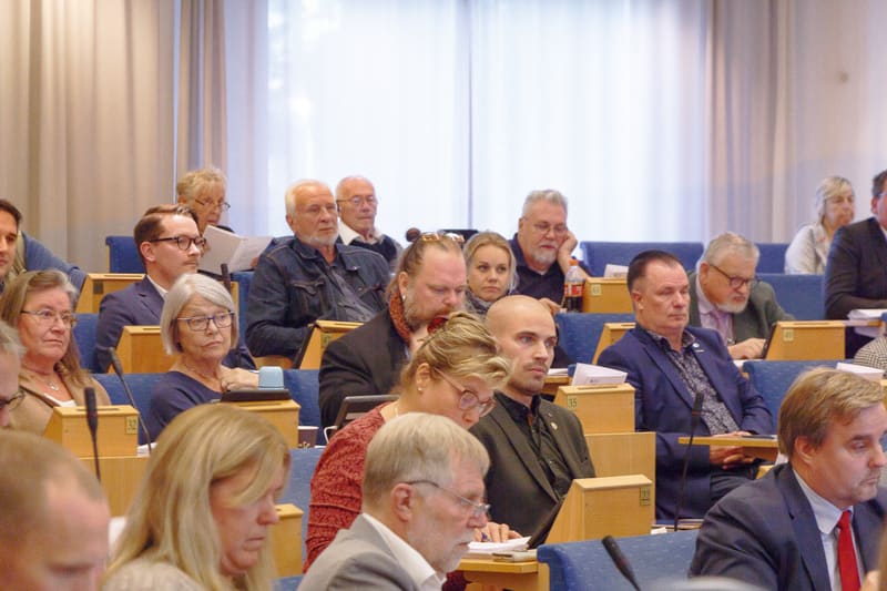Bilden visar en grupp människor som sitter på rader av stolar i en inomhusmiljö. De flesta personerna är vuxna och riktar sin uppmärksamhet framåt, bort från kameran. Mellan stolarna finns små träbänkar eller skrivytor. I förgrunden syns delar av fle