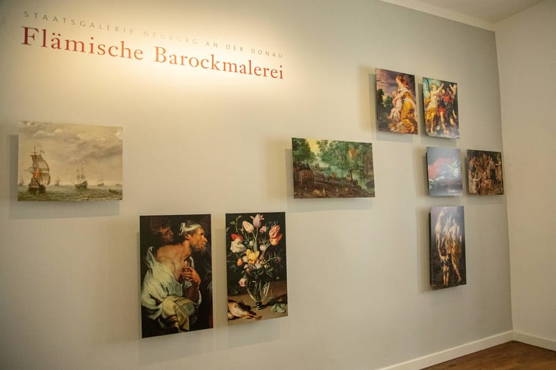 Bilden visar ett inomhusrum med ljusgrå väggar och ett trägolv. På väggen till vänster syns texten "STAATSGALERIE NEUBURG AN DER DONAU FLÄMISCHE BAROCKMALEREI" i rött. Flera tavlor hänger uppe på väggen i varierande storlekar och arrangemang. Till vänster hänger en bild av fartyg på havet. Nedanför den hänger en porträttliknande bild på en man samt en stillbild med en blombukett. På den högra delen av väggen hänger en samling tavlor som föreställer figurer och scener, några med klassiska eller mytologiska motiv.