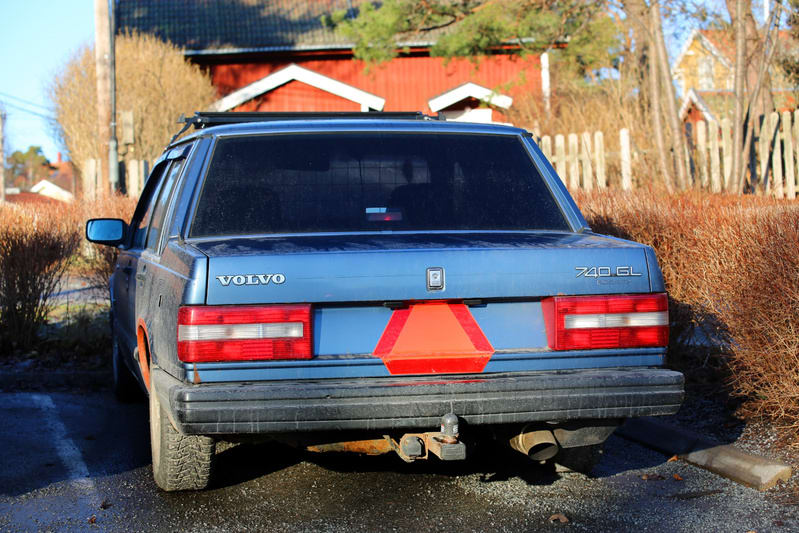Bilden visar bakre delen av en blå Volvo 740 GL sedd bakifrån på en parkering utomhus. Bilen har en varningstriangel (LGF-skylt) fäst under registreringsskylten. Ett takräcke är synligt på bilens tak. I bakgrunden syns delar av en röd byggnad med vita knutar, omgiven av buskar utan löv och ett trästaket. Marken under bilen verkar vara fuktig asfalt eller grus, med en vit parkeringslinje synlig till vänster. Solljus syns på baksidan av bilen och i bakgrunden.