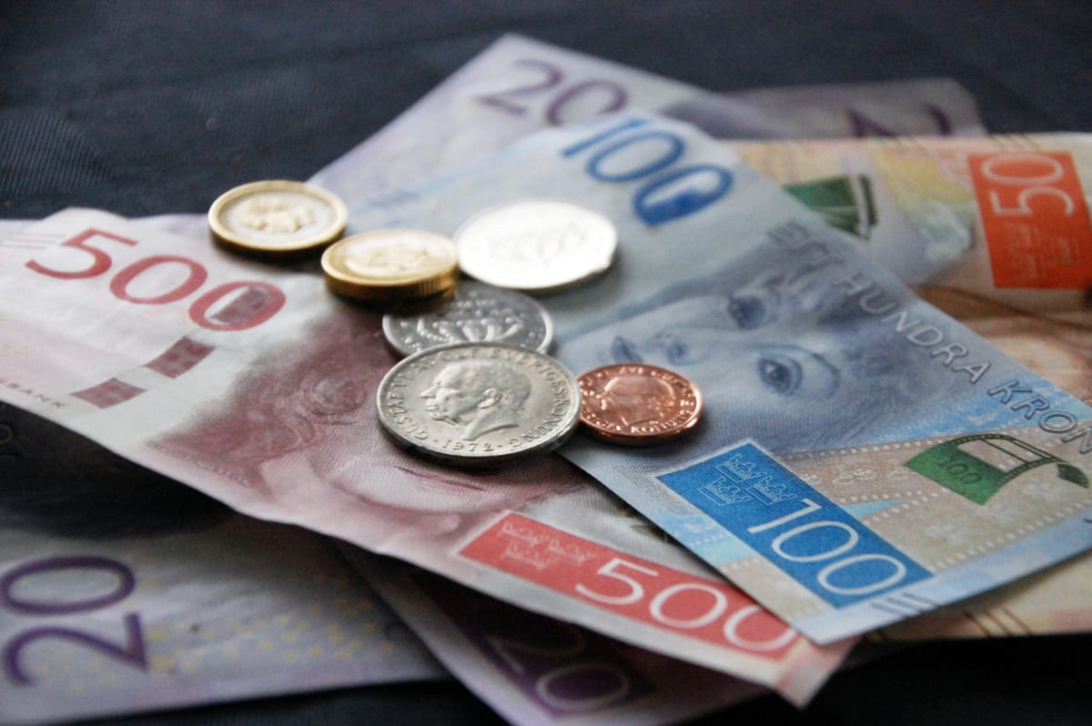 Bilden visar ett arrangemang av svenska sedlar och mynt lagda utspridda på en mörk yta. Bland sedlarna syns valörer som 20, 50, 100 och 500 kronor. Några av sedlarna har avbildningar av ansikten. Flera mynt med olika valörer och metaller (silverfärga