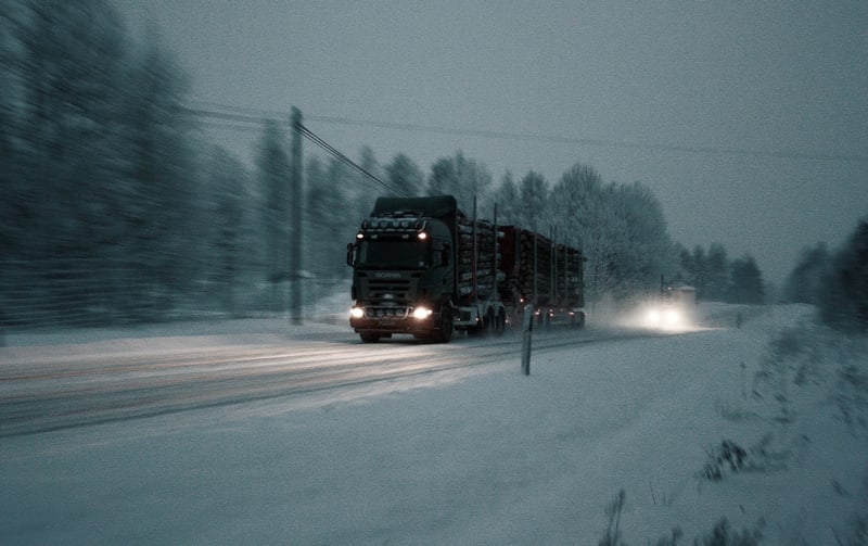 Bilden visar en timmerbil av märket Scania som färdas på en delvis snötäckt väg i skymning eller gryning. Lastbilen har påslagna strålkastare som lyser upp vägen framför. En annan bil med påslagna strålkastare syns i bakgrunden. Kanten av vägen är oc