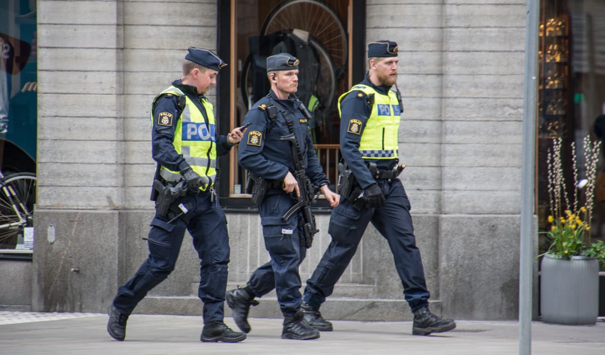 Tre poliser i uniform går på en trottoar längs en byggnad. Polisen längst till vänster bär en gul reflexväst med texten "POLIS" och håller i en mobiltelefon. Polisen i mitten bär också en gul reflexväst med texten "POLIS" och håller i vad som ser ut