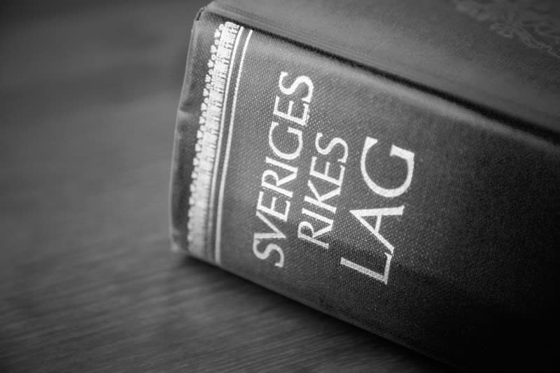 Bilden visar ryggraden på en bok som ligger på ett träbord. Boken är svartvit och texten "SVERIGES RIKES LAG" är tydligt synlig i vita versaler längs med bokryggen. Ryggraden har även ett dekorativt mönster vid dess vänstra kant. Bokens sidor är synl
