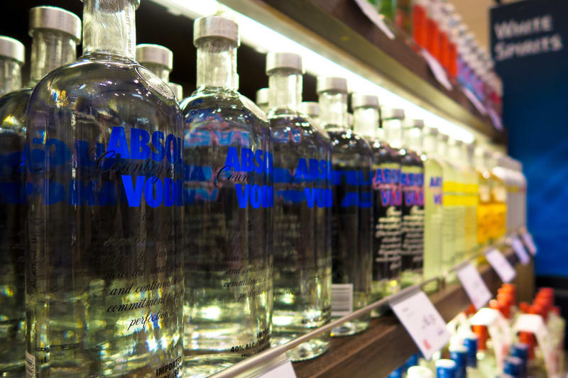 Bilden visar en rad med flaskor, huvudsakligen klara glasflaskor med texten "ABSOLUT COUNTRY OF SWEDEN VODKA" i blått. Dessa står på en trähylla som är upplyst av ett lysrör monterat under hyllan ovanför. Längre bak i bilden syns fler flaskor, några