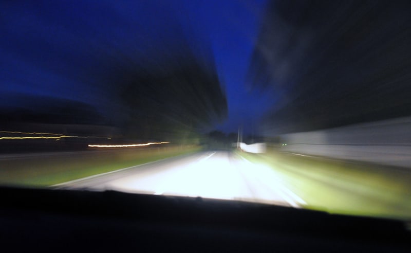 Bilden visar ett väglandskap i mörker, troligen tagen från insidan av ett fordon som rör sig framåt. Mörkblå himmel syns ovanför ett område med suddiga träd eller vegetation. Vägbanan är upplyst av vad som ser ut som strålkastare, vilket skapar ett l