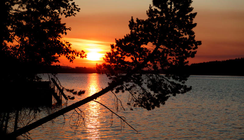 Bilden föreställer en solnedgång över en sjö eller större vattenyta. Solen är placerad i den övre vänstra delen av bilden och lyser starkt med en orange-röd färg, vilket reflekteras på vattenytan som ett brett, skimrande band. I förgrunden syns silhu