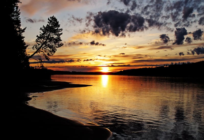 Bilden visar en solnedgång över ett lugnt vattenområde. Solen, som reflekteras starkt i vattnet och skapar ett gyllene sken, är placerad något till vänster om mitten. Horisonten är fylld med varma färger som övergår från gult och orange nära solen ti