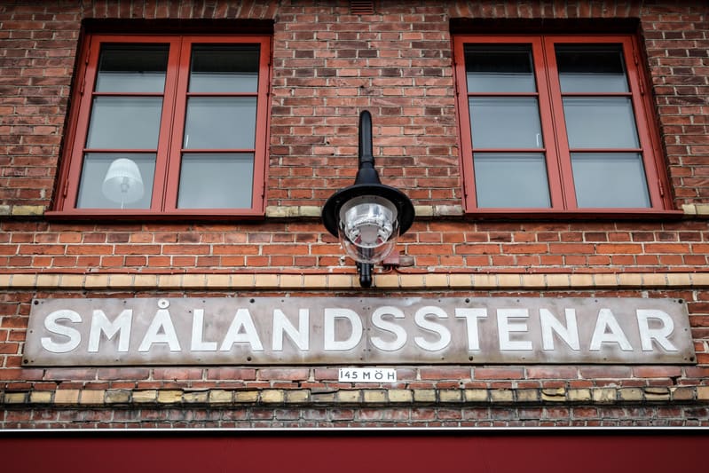 Bilden visar ett utsnitt av en fasad på en tegelbyggnad. Två rektangulära fönster med röda karmar och gråtonat glas syns tydligt i den övre delen av bilden. Mellan fönstren, centrerat vertikalt, sitter en svart lampskylt med en glaskupol, fäst vid te