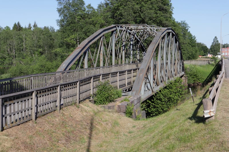 Bilden visar en järnvägsbro med en öppen fackverkskonstruktion, sedd från sidan. Bron har bågformade överliggare och korsande metallbalkar. På båda sidor av bron finns trästaket. Under bron syns grönska och gräs. I bakgrunden till vänster skymtar vat