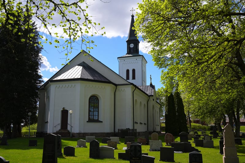 En bild som visar en vit kyrka med ett svart spetsigt klocktorn under en klarblå himmel med några vita moln. Kyrkan har en stor fönsterruta med spröjsning på ena sidan och flera mindre bågformade fönster. I förgrunden syns en kyrkogård med flera grav