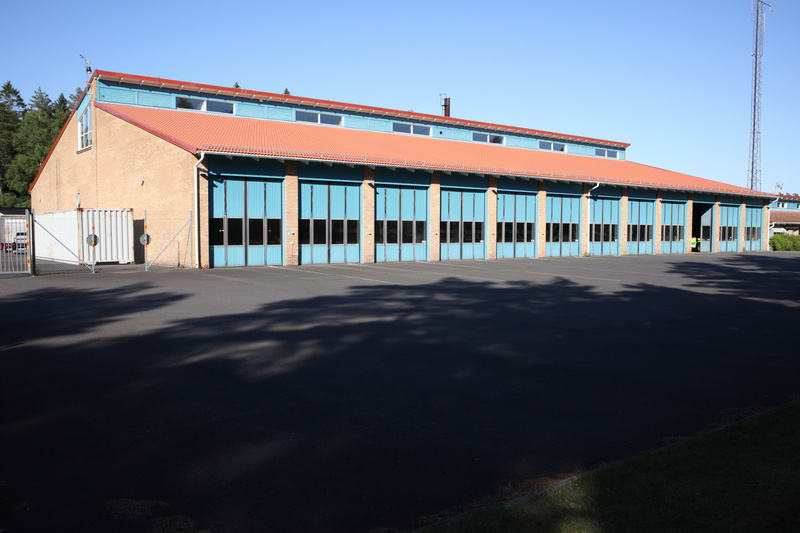 En enplansbyggnad med fasad i ljusgul sten och ett tegelfärgat sadeltak är centralt placerad i bilden. Taket har ett antal takfönster längs långsidorna. Längs byggnadens framsida finns flera garageportar i blått med små, rektangulära fönster. Mellan