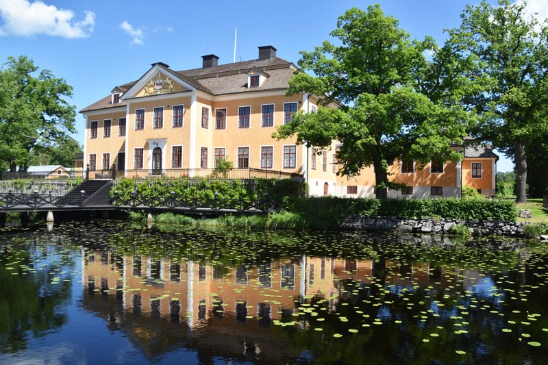 En stor, gul byggnad med många fönster och mörkt tak reflekteras i ett vattenområde. Byggnaden har en barockinspirerad fasad med vita fönsterkarmar och en utsmyckad entré. Framför byggnaden finns ett mörkt träbro som leder upp till en trappa. Längs v