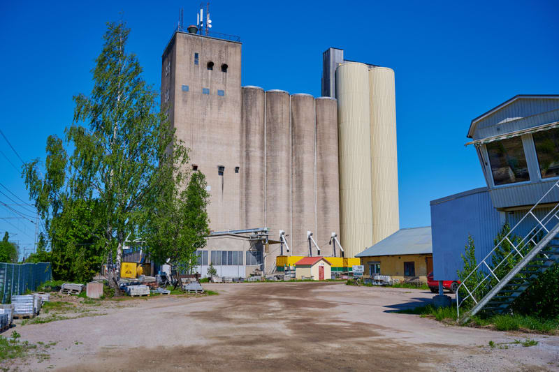 Bilden visar ett industriområde med en stor cementfärgad silo och angränsande byggnader under en klarblå himmel. Siloanläggningen består av flera vertikala cylindriska strukturer, varav några är gråa och andra är ljusgula. En högre, rektangulär byggn