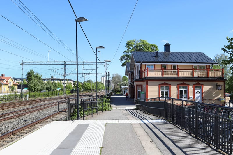 Bilden visar ett landskap med järnvägsspår och byggnader under en klarblå himmel. Till vänster i bilden syns flera parallelle järnvägsspår omgivna av grönska, med järnvägens kontaktledningar och stolpar som spänner över området. Längre bort på vänste