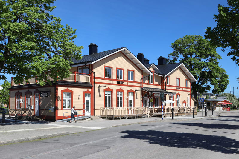 Bilden visar en gulbeige byggnad med röda lister och vitt tak, troligen ett stationshus. Byggnaden har flera fönster och dörrar samt en träveranda. Ovanför några fönster syns texten "TIERP" i stora röda bokstäver. Till vänster om byggnaden finns en d