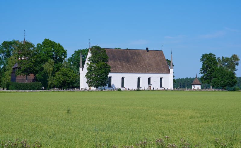 Bilden visar ett landskap med en vit kyrkobyggnad med ett mörkt tak. Bredvid kyrkan, till vänster, står en mörkbrun klockstapel. Mellan kyrkan och klockstapeln finns en mur som omger en kyrkogård med ett flertal gravstenar. I bakgrunden syns många gr