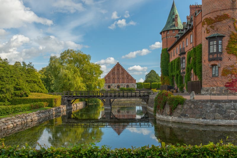 Bilden visar ett stort tegelbyggnadskomplex med en vallgrav framför. Till höger syns en stor tegelbyggnad, antagligen en del av ett slott eller en borg, med ett spetsigt grönt taktorn och täckt av klätterväxter, både gröna och rödaktiga. Byggnaden ha