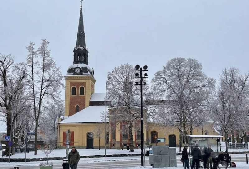 Bilden visar en gatuscen på vintern med en kyrka i bakgrunden. Kyrkan har en hög, mörk spira med en klocka högst upp och är byggd i tegel och gulmålad puts med snö på taket. Flera träd med frostiga, snötäckta grenar står framför och runt kyrkan. I fö