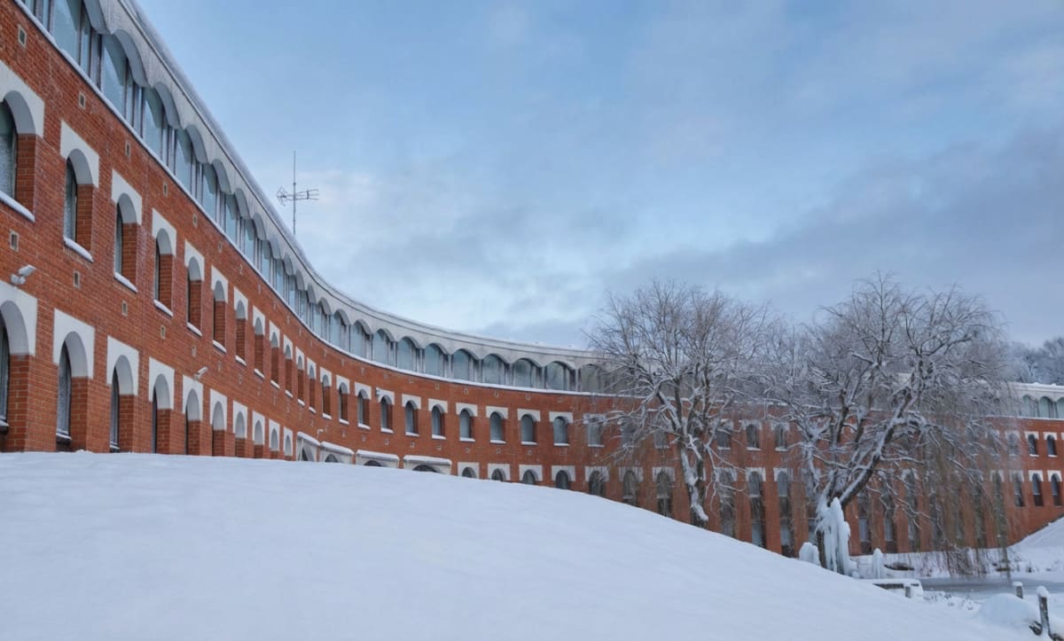 Bilden visar en röd tegelbyggnad med många valvformade fönster. Byggnaden har en böjd form och är belägen i ett vinterlandskap täckt av snö. På en snötäckt kulle i förgrunden finns det kala träd med snö på grenarna. En del av träden har istappar. Bakom träden syns en frusen vattenyta. Himlen är delvis mulen med inslag av blått. Byggnadens taklinje har också valvformade fönster och är täckt med snö och is. På taket syns en telekommunikationsantenn. Den vita snön kontrasterar mot den röda tegelfasaden.